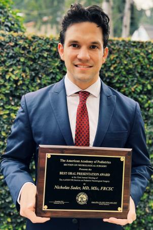 Nicholas Sader, MD, MSc, FRCSC, smiles while standing outdoors holding a dark wood rectangular plaque with gold lettering.
