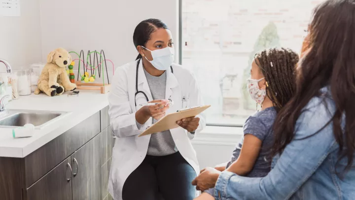A female doctor with medium-dark skin tone sits in an exam room and speaks to a child sitting on a woman's lap. All are wearing masks.