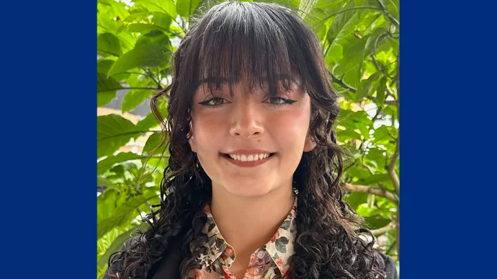 Headshot of a smiling young woman with medium skin tone and dark hair wearing a floral print blouse against an outdoor background