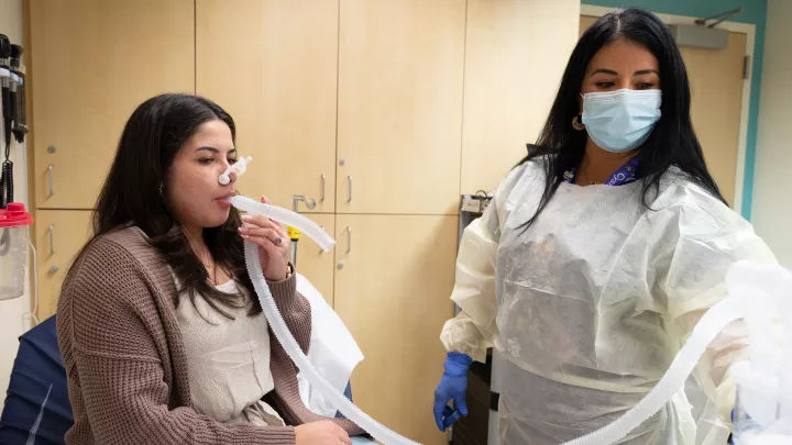 Casually dressed female patient with medium skin tone and dark hair blows into a long, clear plastic tube connected to a pulmonary testing device and monitored by a female nurse with medium skin tone and dark hair wearing surgical mask, gown and gloves