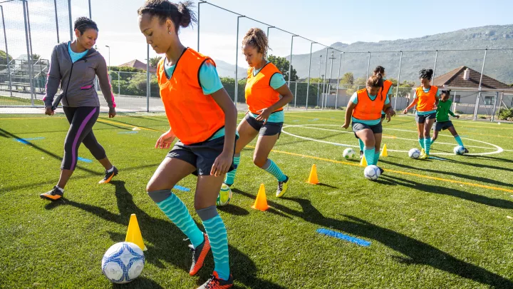 Six teen girls dribble soccer balls around cones, while a female coach watches.