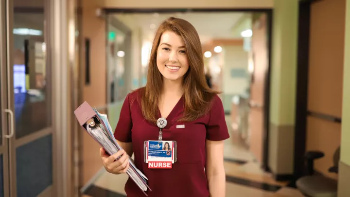 Nurse with light skin tone and auburn hair wearing a maroon nurses uniform and holding a binder smiles as she stands in a hospital corridor