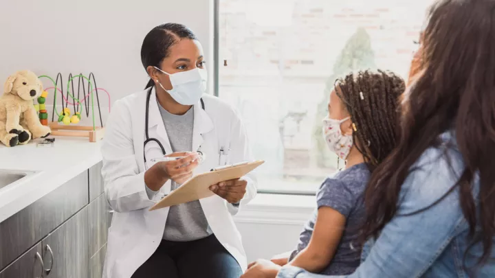A masked health care provider with medium skin tone and dark hair takes information from a young mother and daughter in a medical office