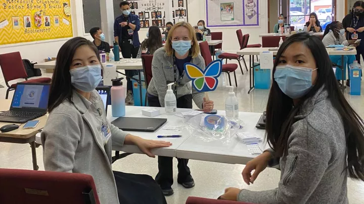 A seated medium-light skin toned woman, a standing light skin-toned woman holding a butterfly sign, and a seated medium skin-toned woman, all wearing surgical masks, smile at the camera while nurses administer vaccinations behind them