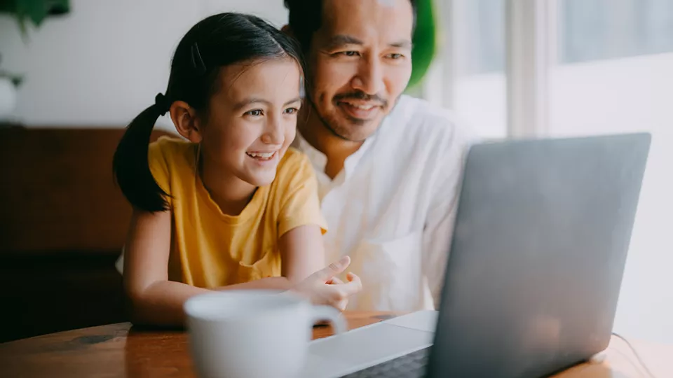 Happy father and young daughter smile at laptop screen in their home
