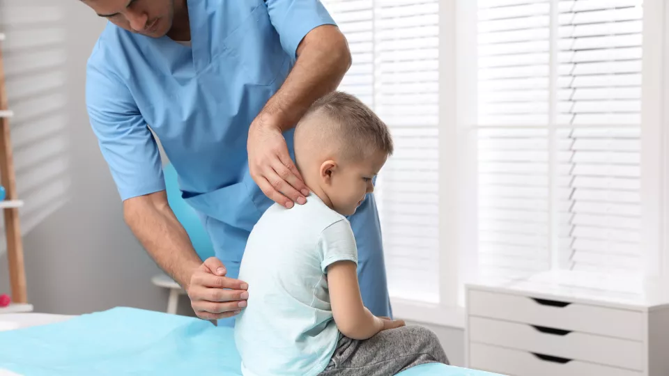 A doctor in blue scrubs checks a young boy’s back during an exam. The boy wears a light T-shirt and is sitting on an exam table
