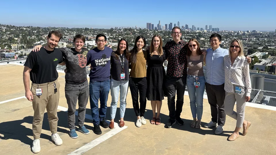 A group of 10 casually dressed doctors stand shoulder to shoulder on Children's Hospital Los Angeles helipad with the downtown Los Angeles skyline in the background on a bright, sunny day