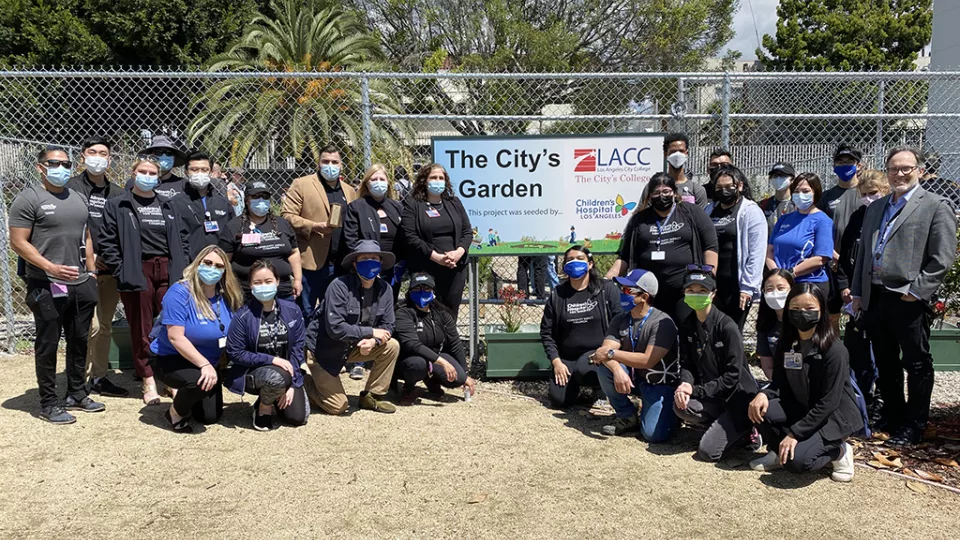 A group of 27 people, most in black shirts or jackets with the CHLA logo, pose and smile with a sign that says: The City's Garden. The Sign also shows the CHLA and Los Angeles Community College logos.