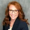 Headshot of a smiling woman with light skin and red hair wearing tortoise shell glasses and a dark suit jacket