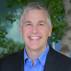 Headshot of a smiling man with light skin tone and grey hair wearing a blue, open-collar dress shirt under a dark suit jacket against a blurred outdoor background