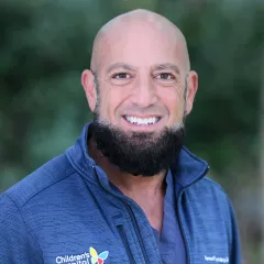 Headshot of a smiling man with light skin tone, shaved head and thick dark beard wearing a dark blue full-zip against a blurred outdoor background