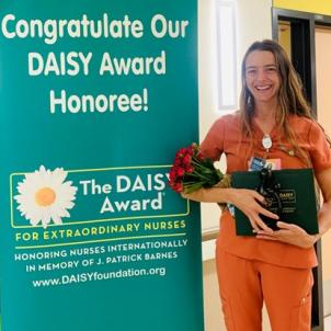 Smiling woman with light skin tone and long brown hair wearing orange nurse scrubs and holding bouquet of red flowers and her DAISY Award poses in hospital corridor next to DAISY Award poster
