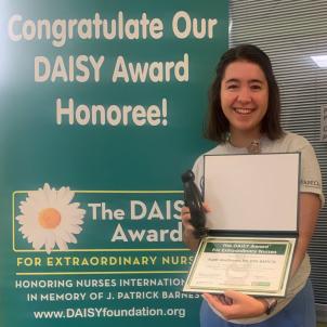 Smiling female nurse with light skin tone and dark, shoulder-length hair poses with their DAISY Award next to congratulatory poster