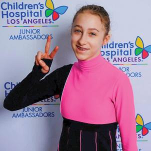 A smiling teenage girl with light skin tone and blonde hair wearing a pink and black top flashes the peace sign in front of a Children's Hospital Los Angeles backdrop