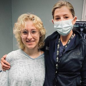 Smiling young woman with light skin tone and blonde hair wearing a hospital gown stands with a female physician with light skin tone and light brown hearing wearing a surgical mask