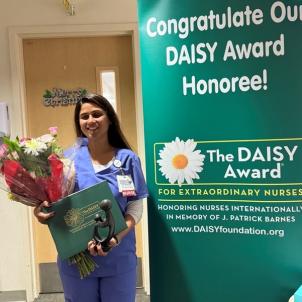 Smiling female nurse with medium skin tone and long brown hair holds bouquet of flowers and award statue next to congratulatory poster in hospital setting