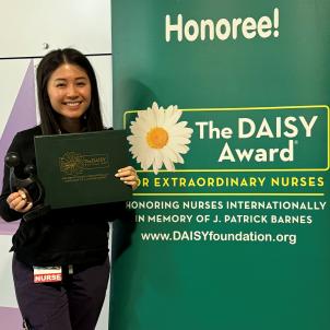 A young woman with medium skin tone and straight dark hair smiles as she poses with her DAISY Award statue next to a DAISY Award poster