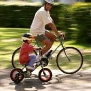 An adult rides a bike next to a small child on a bike with training wheels. Both are wearing helmets.