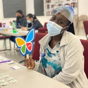 A dark skin-toned woman wearing scrubs, a surgical mask and a face shield sits, holding a CHLA butterfly logo sign and smiling