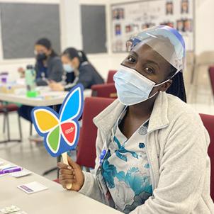 A dark skin-toned woman wearing scrubs, a surgical mask and a face shield sits, holding a CHLA butterfly logo sign and smiling