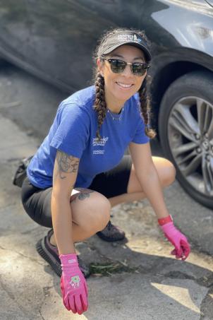 Medium skin toned woman wearing sunglasses, protective gloves and CHLA branded visor and t-shirt smiles at the camera as they clean sidewalk