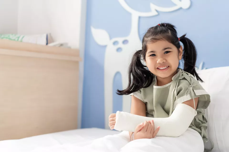 A girl with medium skin tone sits in a hospital bed, smiling and cradling her casted arm. She wears pigtails and a hospital gown.