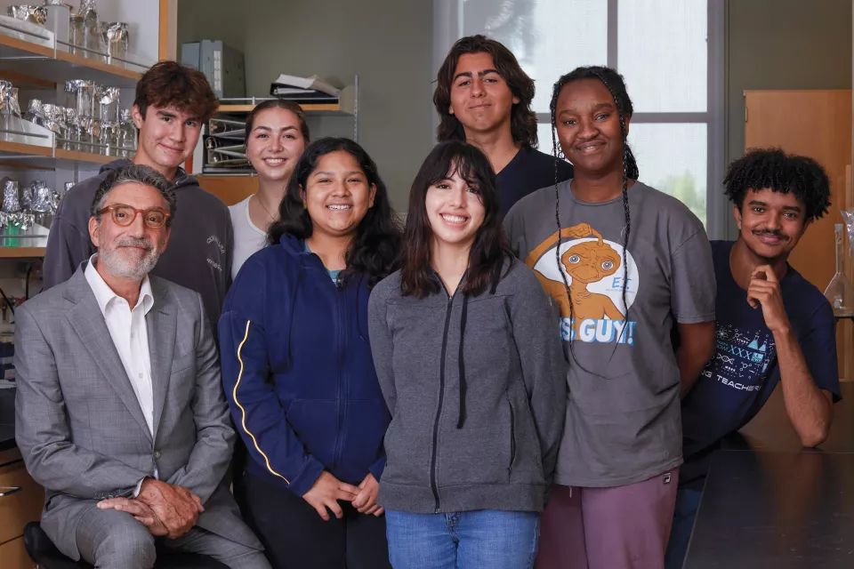 Chuck Lorre, a man with light skin and grey hair, wearing a white shirt and grey suit sits with seven casually dressed high school students in a laboratory setting