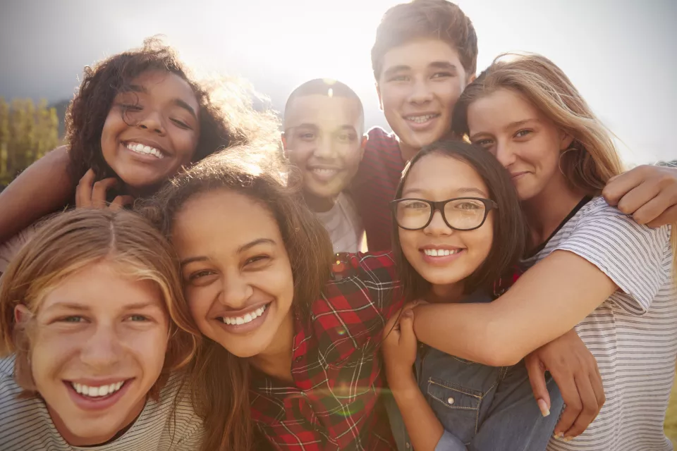 Closeup of seven, smiling, casually dressed teenagers with light, medium and dark skin tones in a group hug on a bright day outdoors