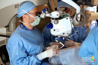 Two men in surgical gowns, head coverings and masks look through a device while performing a procedure