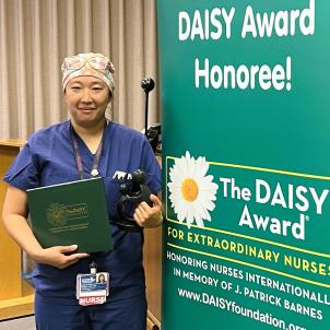 Female nurse with medium skin tone and wearing blue scrubs smiles as they pose with their DAISY Award next to a DAISY Award poster
