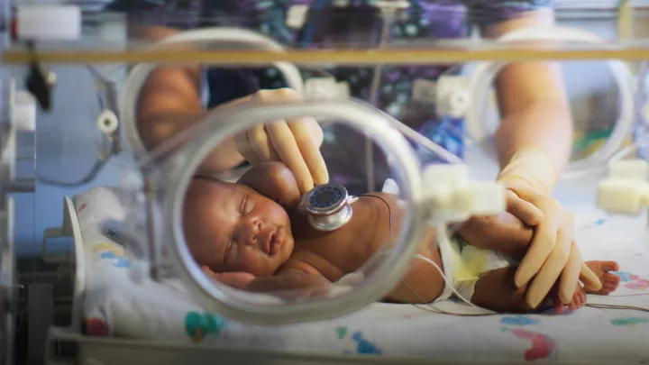 A woman with light skin tone holds a stethoscope on the chest of a medium-dark skin toned infant in an incubator.