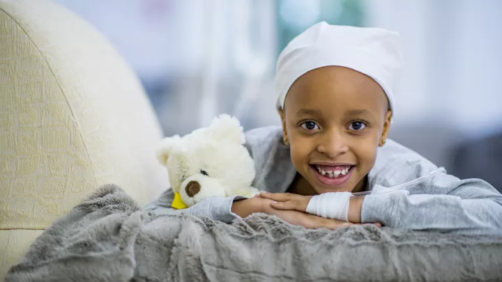 A little girl with medium skin tone lays on a cushion, smiling. She holds a teddy bear and wears a headscarf. A tube is bandaged to the back of her hand.