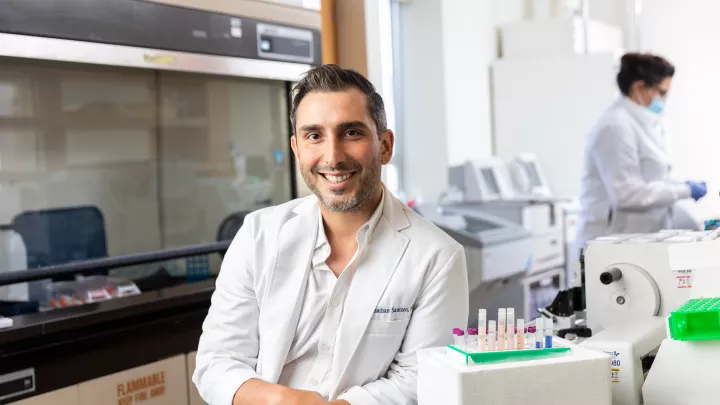 A man with medium skin tone wearing a lab coat leans on a research laboratory bench and smiles at the camera.