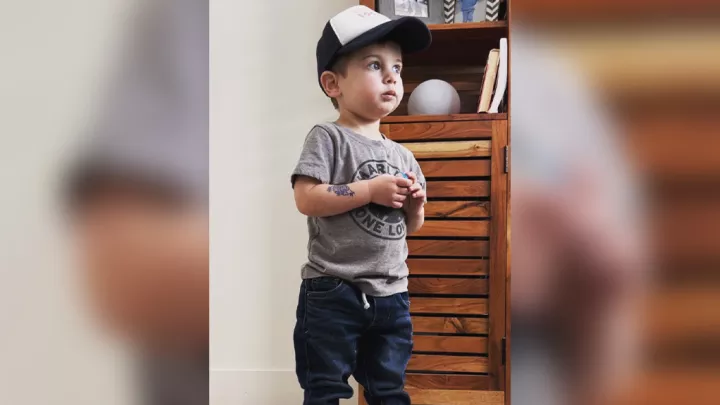 Little boy Kai stands next to a bookcase wearing a baseball cap, grey t-shirt and blue jeans