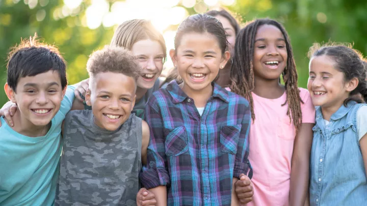 A group of six smiling children pose for the camera arm-in-arm in the afternoon sunshine