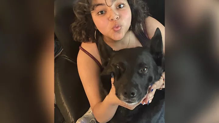 Kairi, a medium skin tone teenager, sits in a black leather chair and blows a kiss toward the camera while holding her black dog in her lap