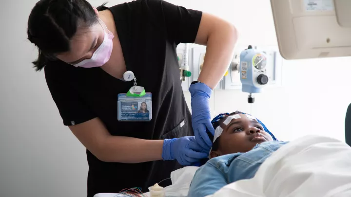 A medium-light skin toned woman in black scrubs and a surgical mask tapes wires to the head of a dark skin toned girl lying on a hospital bed.