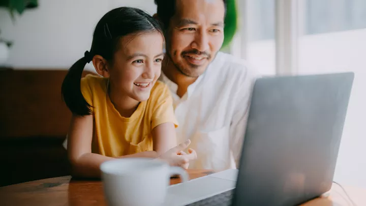 Happy father and young daughter smile at laptop screen in their home