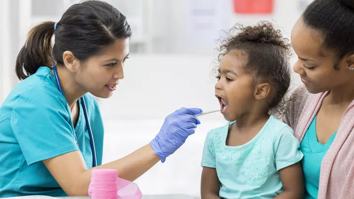 Pediatrician uses a tongue depressor to examine a young female patient's throat as she sits on her mother's lap