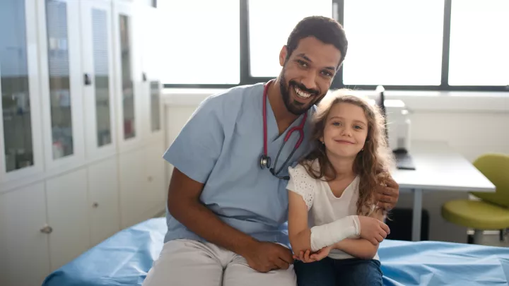 A medium-dark skin toned man in scrubs with a stethscope poses next to a light skin toned girl holding her bandaged arm. Both smile. 