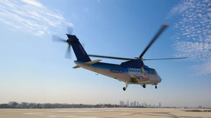 A Sikorsky S-76 helicopter with the Children's Hospital Los Angeles logo hovers above a helipad. The sky is blue and streaked with clouds and downtown L.A. is in the background.