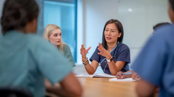 A medium skin toned woman in scrubs and a stethoscope talks to colleagues sitting around a table. 