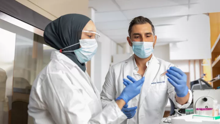 A woman with medium-light skin tone wearing a hijab shows a sample tube to a man with medium skin tone. Both wear lab coats and face masks.