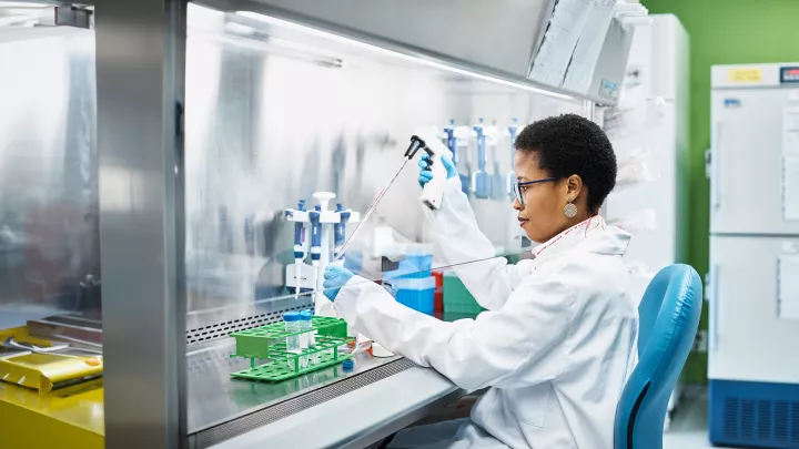 A woman with medium dark skin tone in a lab coat uses a pipetting device in a laboratory fume hood. 