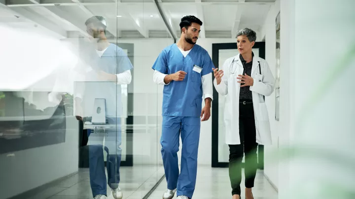 A medium-light skin toned woman with short gray hair wearing a lab coat and stethoscope talks to a medium skin toned man in blue scrubs as they walk. 