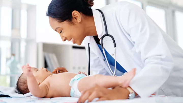 A medium skin toned woman in a lab coat and stethoscope smiles and examines a medium-light skin toned baby in a diaper. 
