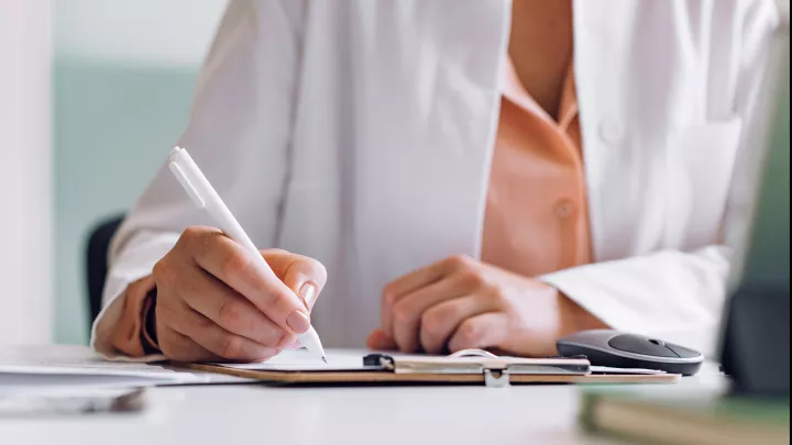 A view of the hands of a woman in a lab coat writing on a medical chart. 