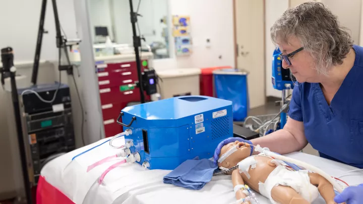 A light skin toned woman in blue scrubs leans over a simulated model of an infant laying on a hospital bed. 