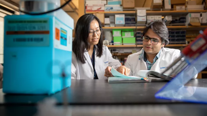 A medium skin toned woman points at a book that a medium skin toned man is looking at in a research laboratory. Both wear lab coats. 