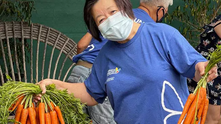Middle aged woman wearing a blue CHLA t-shirt and a medical mask holds several bunches of carrots 